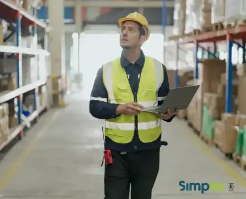 Trabajador con casco y chaleco reflectante utiliza una computadora portátil en un depósito.