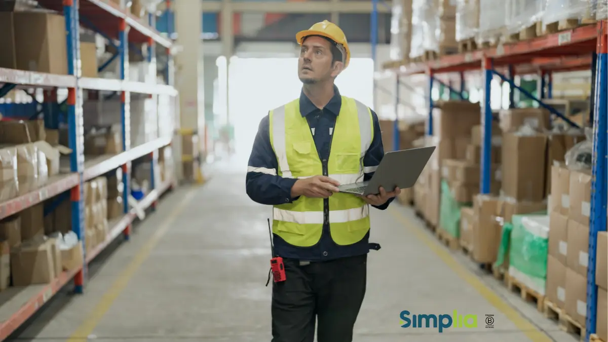 Trabajador con casco y chaleco reflectante utiliza una computadora portátil en un depósito.
